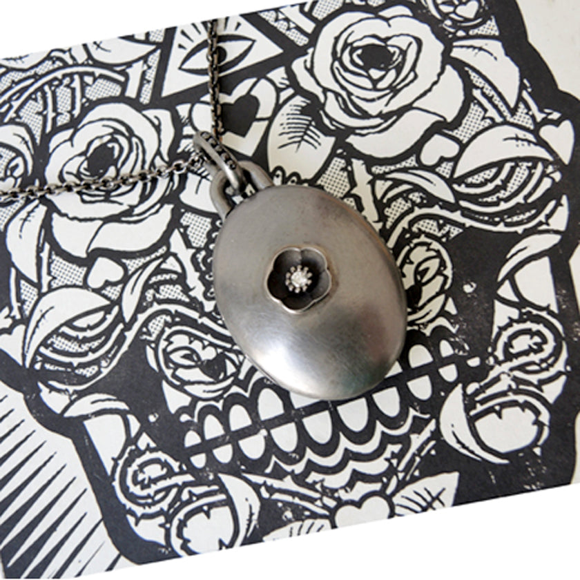 Blackened silver locket with a black flower-shaped center with a diamond on a black and white patterned card.