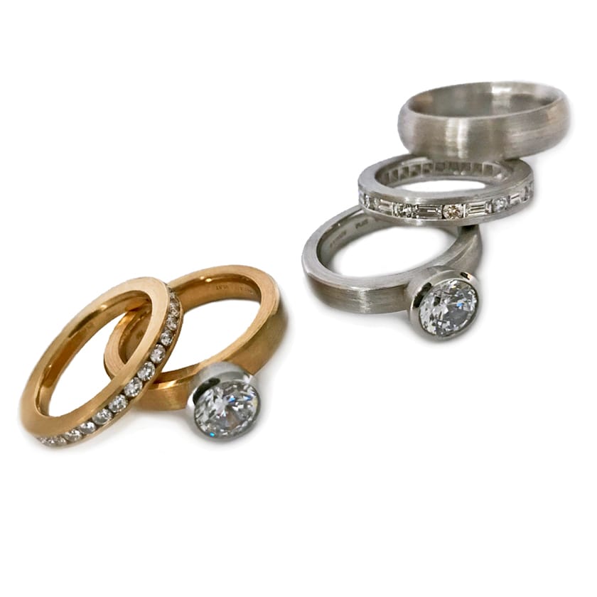 Solitaire bezel set engagement rings in gold and platinum with channel set wedding bands and a plain matte wedding band for a man.